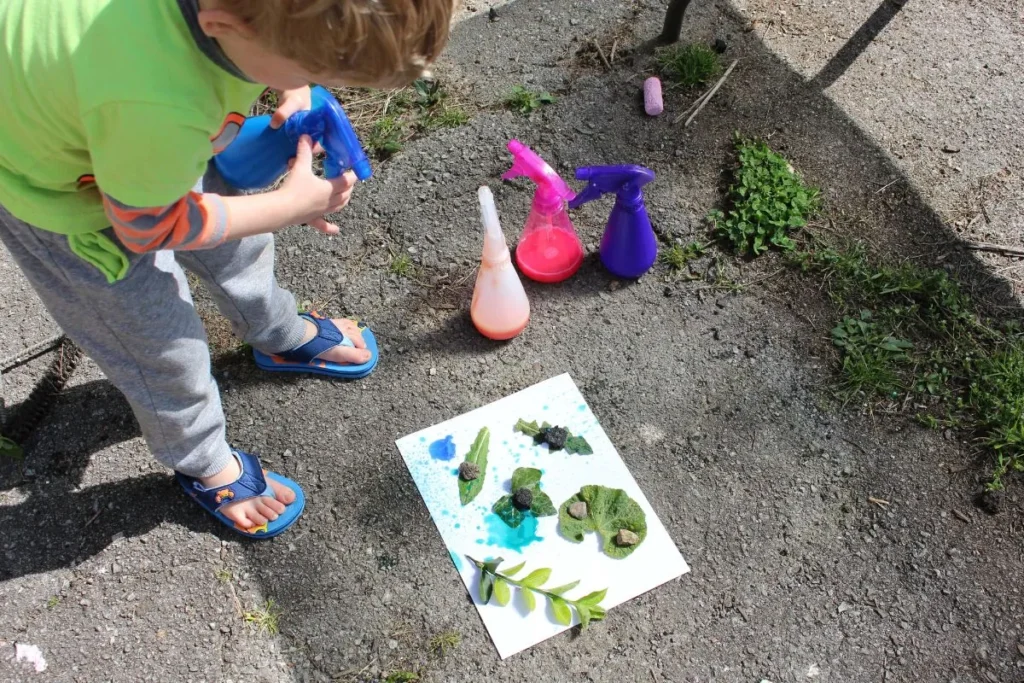 A young child creating spray bottle art by spraying paint over a paper covered with leaves.